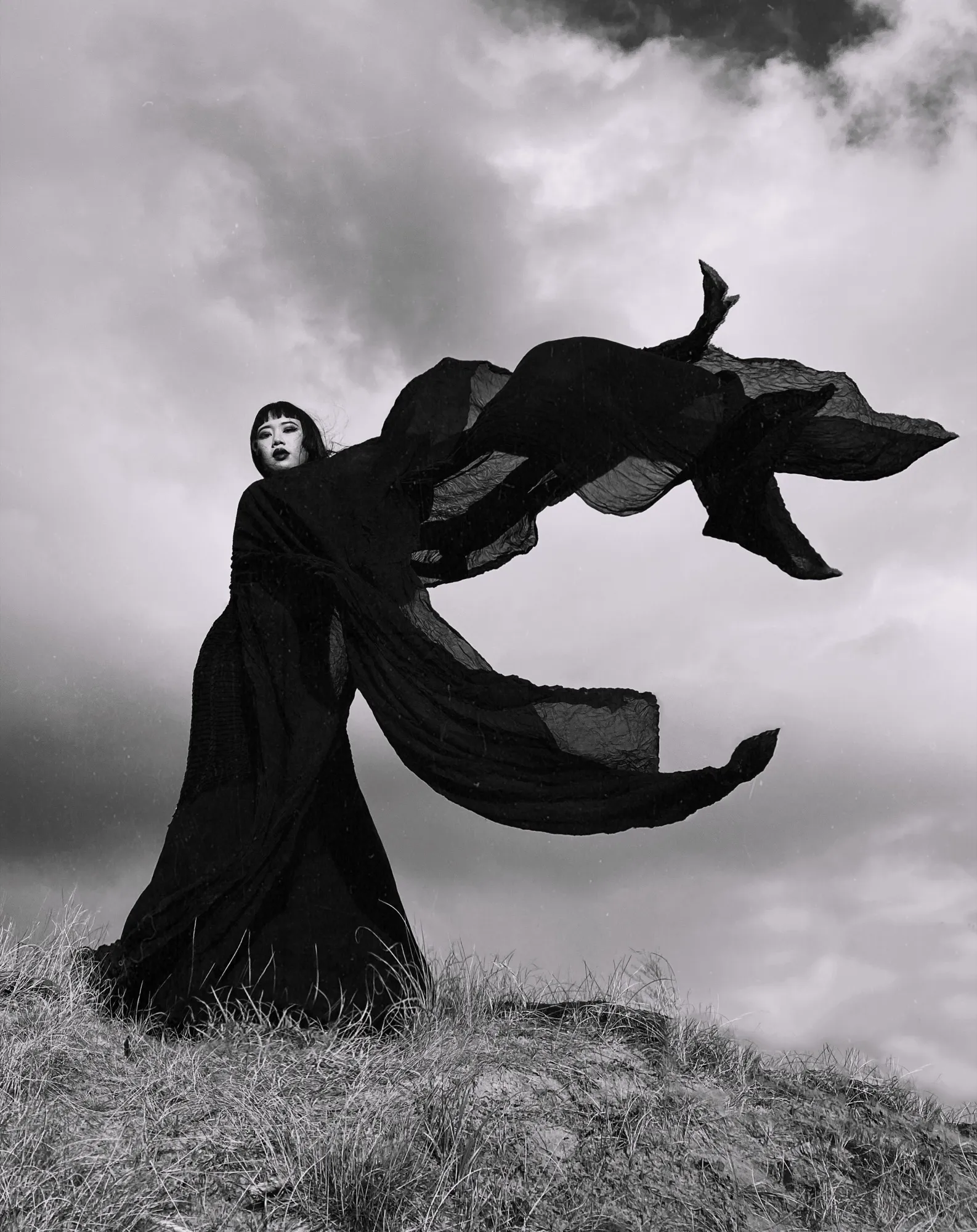 Black-and-white photograph: a figure on a windswept hill, black fabric billowing like wings against the clouds